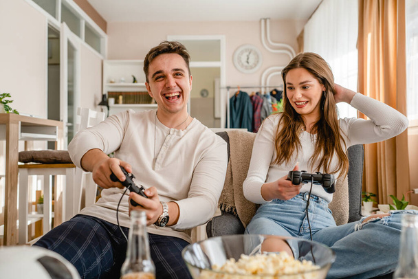 twee Kaukasische mensen zitten thuis spelen video game console met plezier jong stel man en vrouw vriend en vriendin samen tijd doorbrengen met joystick controller glimlachen vooraanzicht - Foto, afbeelding