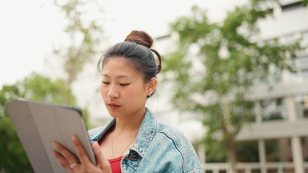 Estudiante bastante asiática usando tableta digital durante las vacaciones de estudio al aire libre. Sonriendo chica japonesa haciendo la tarea en la tableta PC sentado en la calle - Foto, Imagen