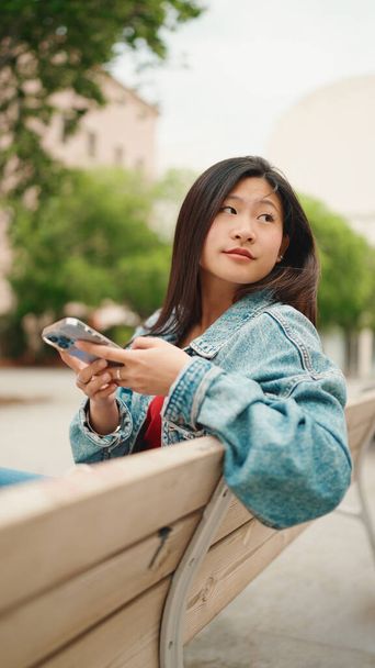Porträt eines langhaarigen asiatischen Mädchens, das lässig gekleidet auf der Bank sitzt und auf der Straße ihr Smartphone kontrolliert - Foto, Bild