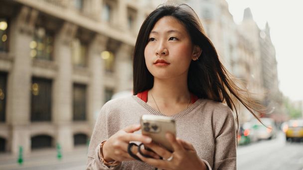 Retrato de asiática de pelo largo chica buscando pensativo vestido casual mensajes de texto con amigos en la calle. Mujer joven usando móvil al aire libre - Foto, imagen