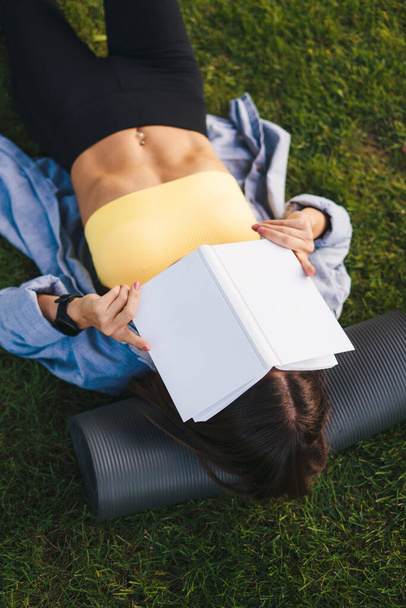 Вид сверху на женщину, одетую в хиджаб, лежащую на траве, упирающуюся головой в коврик для йоги и закрывающую лицо книгой. Дистанционное обучение, зарабатывание и - Фото, изображение