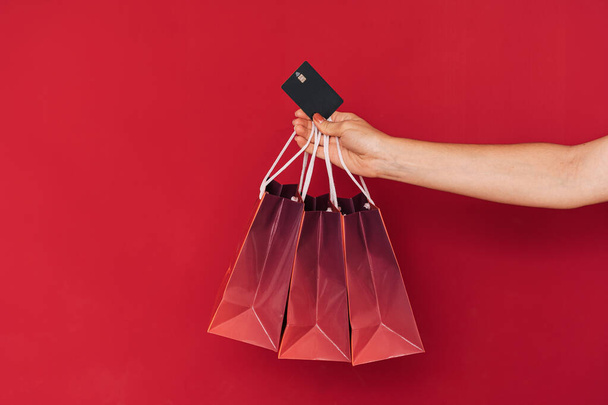 Main tenant trois sacs en papier et une carte de crédit devant un fond rouge. - Photo, image