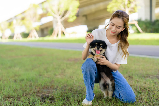 Szczęśliwa młoda Azjatka bawiąca się i siedząca na trawie w parku ze swoim psem. Koncepcja miłośnika zwierząt - Zdjęcie, obraz
