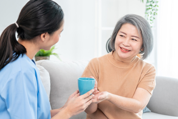 Female care taker serving her contented senior patient with a cup of coffee at home, smiling to each other. Medical care for pensioners, Home health care service. - Foto, Imagem