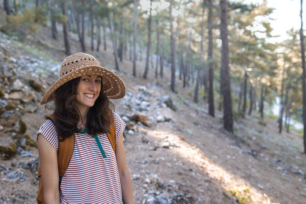 Retrato de uma mulher com uma mochila e um chapéu de palha na floresta, uma menina sorridente em uma caminhada, trekking verão, mulher feliz desfrutando de seu passeio na floresta - Foto, Imagem