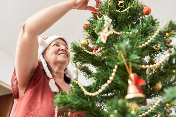 Felice donna latina anziana sorridente mentre decorava il suo albero di Natale a casa, indossando un berretto rosso di Babbo Natale con trecce bianche. La gioia delle vacanze. - Foto, immagini