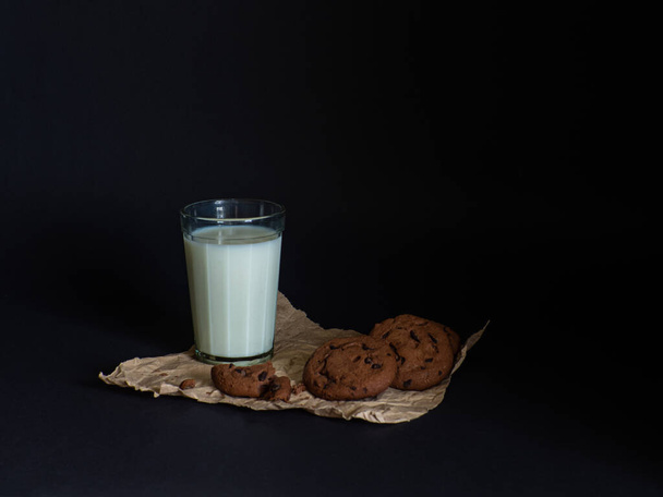 Cookies made from cocoa and with pieces of chocolate inside and glass of milk on a baking paper on a black background. Breakfast eating. Copy space - Foto, imagen