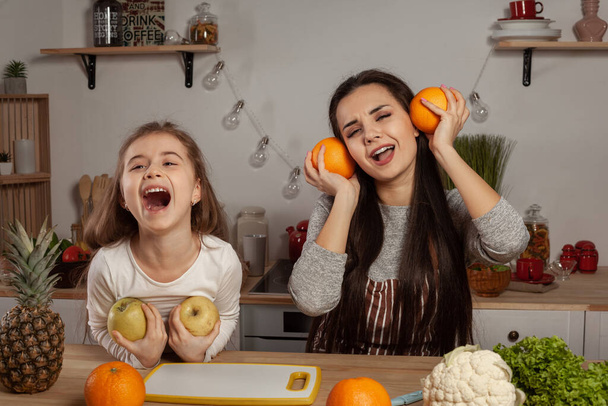 Happy loving family are cooking together. Elegant mom and her kid are doing a fruit cutting and fooling around at the kitchen, against a white wall with shelves and bulbs on it. Homemade food and - Fotoğraf, Görsel