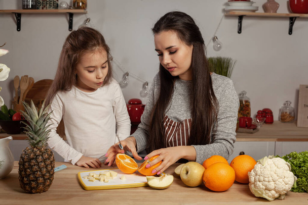 Happy loving family are cooking together. Alluring mom and her kid are doing a fruit cutting and having fun at the kitchen, against a white wall with shelves and bulbs on it. Homemade food and little - Photo, Image