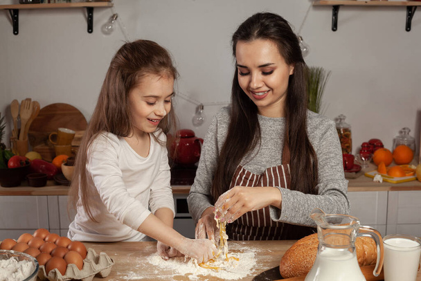 Happy loving family are preparing pastries together. Alluring mom and her kid are adding an egg to the dough and having fun at the kitchen, against a white wall with shelves and bulbs on it. Homemade - Фото, зображення