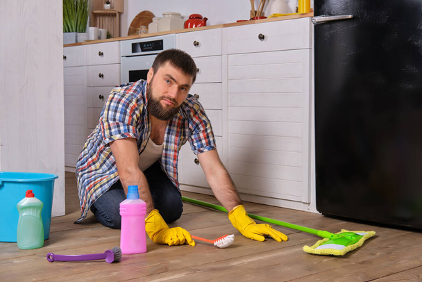 Концепция Housecleaning. Белый красивый молодой бородатый мужчина в клетчатой рубашке сидит на полу своей кухни и пытается почистить ее всеми своими моющими средствами, тряпками, мочалками и щетками
 - Фото, изображение