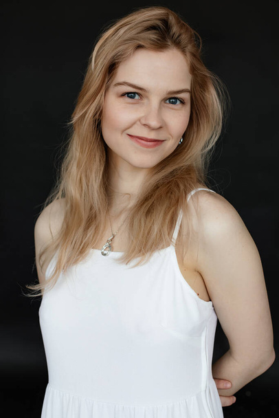 Vertical portrait of positive, smiling and cheerful young blonde long haired woman with perfect shiny skin in white top and necklace look at camera on black isolated background. Natural beauty - 写真・画像