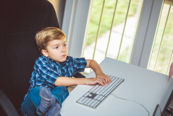 Pieni poika kirjoittaa tietokoneen näppäimistöllä rentoutuessaan jalat pöydällä kotona.  - Valokuva, kuva