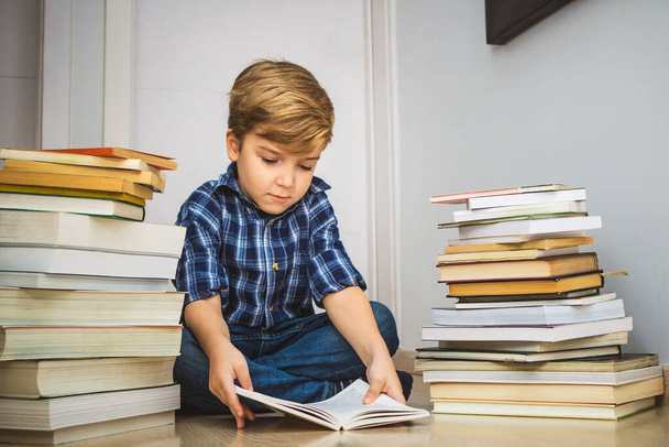 Kleiner Junge liest, während er auf dem Boden zwischen Bücherhaufen sitzt.  - Foto, Bild