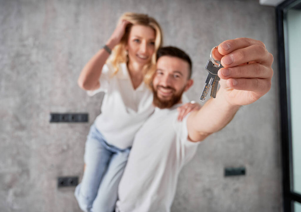 Fröhlicher Mann hält Frau und lächelt, während er Wohnungsschlüssel vorzeigt. Glückliche Immobilienkäufer mit Schlüsseln aus dem neuen Zuhause. Männerhand mit Schlüsseln im Fokus. - Foto, Bild