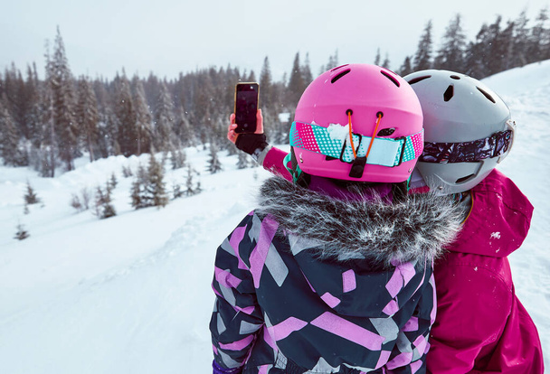 Back view of mother and daughter in ski equipment together taking selfies or video call in the mountains on ski resort at sunny winter day, travel vacation, or network coverage and connection concept. - Photo, image