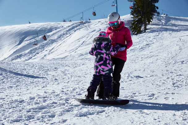 A mãe ensina sua filha a snowboard. Mulher ajudando e apoiar criança passeio de snowboard na estância de esqui no dia ensolarado de inverno, férias de viagem, paisagem montanhas fundo. - Foto, Imagem