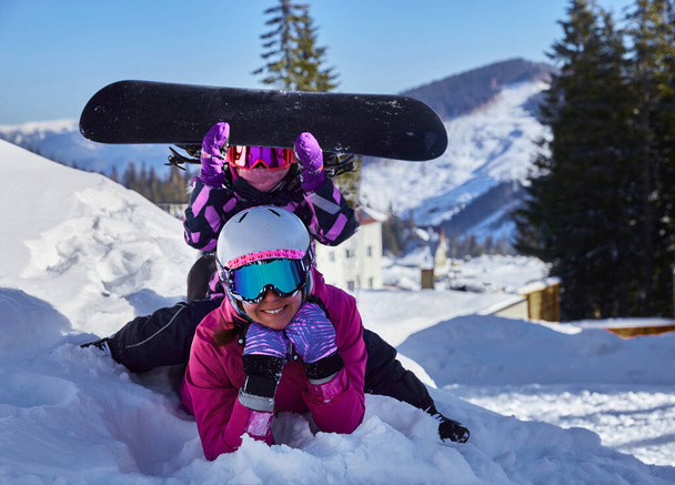 Mãe e filha no equipamento de esqui juntos deitado na neve desfrutando de inverno estão se divertindo na estância de esqui. Família feliz brincando na neve com um snowboard - Foto, Imagem