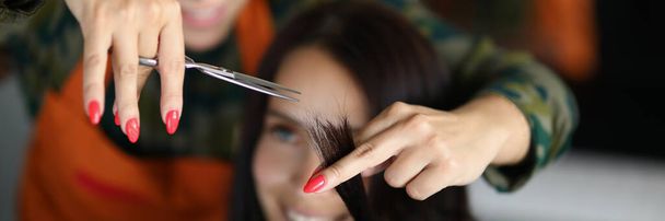Портрет концентрированного профессионального парикмахера держит прядь волос и стрижет ножницами. Улыбающаяся клиентка сидит в кресле хозяина. Концепция здоровья - Фото, изображение