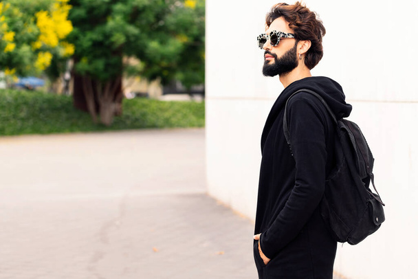 beautiful young man with backpack and fashionable sunglasses walking by the city, concept of urban lifestyle and stylish clothing, copy space for text - Photo, Image