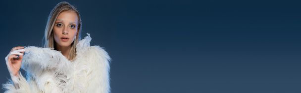 young woman with winter makeup posing in faux fur jacket touching feather on dark blue, banner - Photo, Image