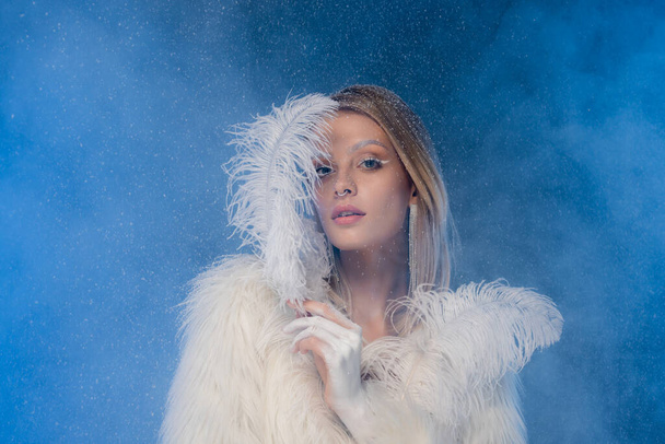 blonde woman with winter makeup in faux fur jacket holding white feather under falling snow on dark blue - Foto, afbeelding