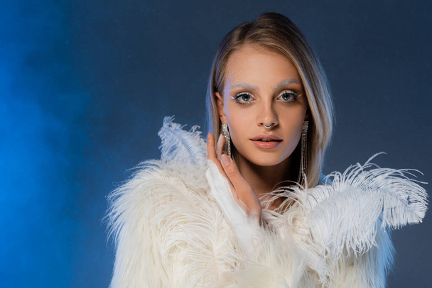 young pierced woman in faux fur jacket with white feathers posing on dark blue background - Photo, Image