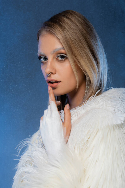 blonde woman with winter makeup and frozen eyebrows touching lip on dark blue  - Foto, Imagen