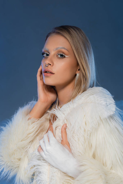 pierced woman with winter makeup posing in white faux fur jacket on dark blue  - Photo, Image
