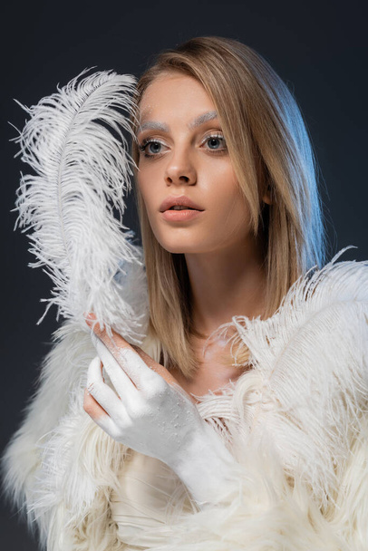 blonde young woman with winter makeup posing in white faux fur jacket holding feather isolated on blue - Foto, imagen