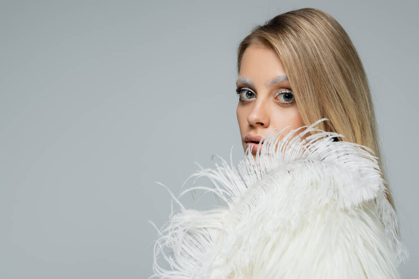 young woman in faux fur jacket looking at camera near white feather isolated on grey - Φωτογραφία, εικόνα