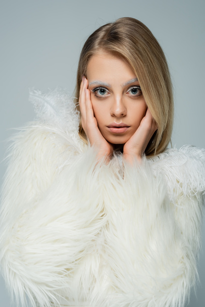 blonde young woman in stylish faux fur jacket with white feathers looking at camera isolated on grey - Photo, Image