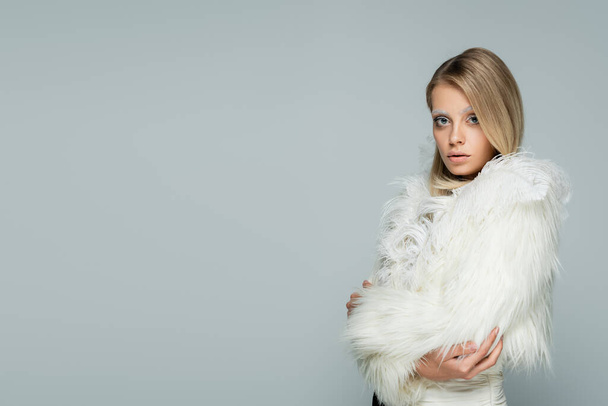 молодая модель в стильной искусственной меховой куртке с белыми перьями, смотрящая в камеру, позируя изолированно на сером - Фото, изображение