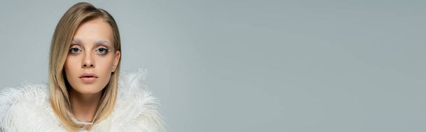young model in stylish faux fur jacket with white feathers looking at camera isolated on grey, banner - Foto, imagen