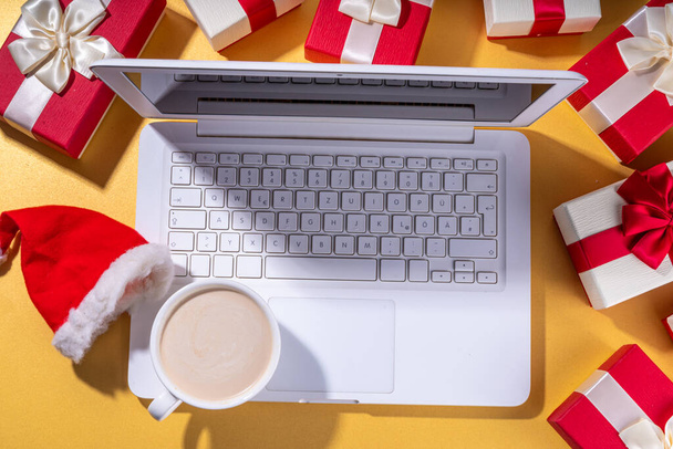 Kerstverkoop, voorbereiding op vakantie concept. Kerstman handen met laptop, warme chocolademelk latte cup, geschenkdozen, plat lag op gouden achtergrond. Maken van verlanglijst, voorbereiding geschenken voor Kerstmis. - Foto, afbeelding