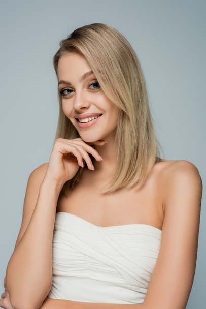 happy blonde woman with bare shoulders and natural makeup looking at camera isolated on grey - Foto, immagini