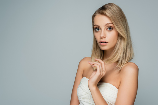 blonde woman with bare shoulders and natural makeup looking at camera isolated on grey - 写真・画像