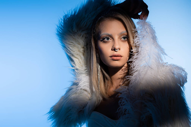 pierced young woman with winter makeup and white eyebrows posing with feather on blue - Foto, Imagem