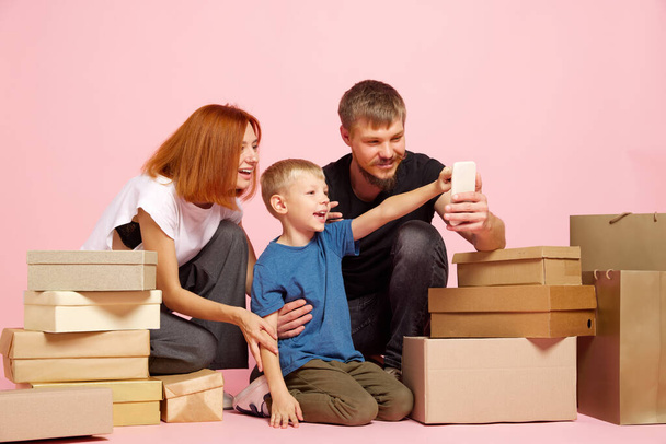 Happy mother, father and son open cardboard boxes, receive parcels unpack after online shopping over pink studio background. Unbox package, post shipping delivery and big sales day concept - Foto, Imagem