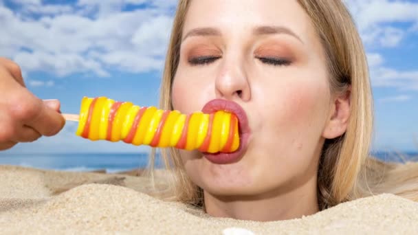 A pretty woman buried up to her head on a beach licking a lollipop held by another hand - Footage, Video