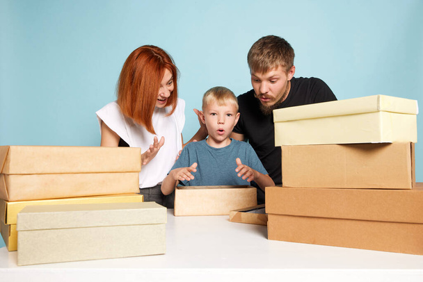 Unpacking. Friendly family, parents and child open cardboard boxes, receive parcels unpack after online shopping over blue background. Unbox package, post shipping delivery and big sales day concept - Photo, image
