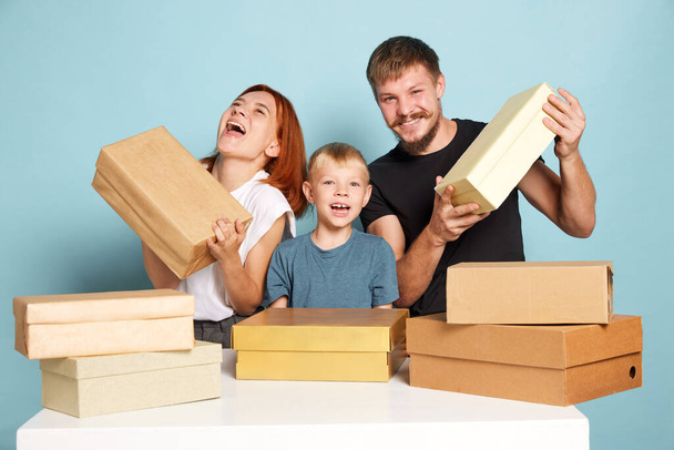 Unpacking. Friendly family, parents and child open cardboard boxes, receive parcels unpack after online shopping over blue background. Unbox package, post shipping delivery and big sales day concept - Foto, Imagen