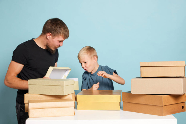 Looking with curiosity. Young father and child open cardboard boxes, receive parcels unpack after online shopping over blue background. Unbox package, post shipping delivery and big sales day concept - 写真・画像
