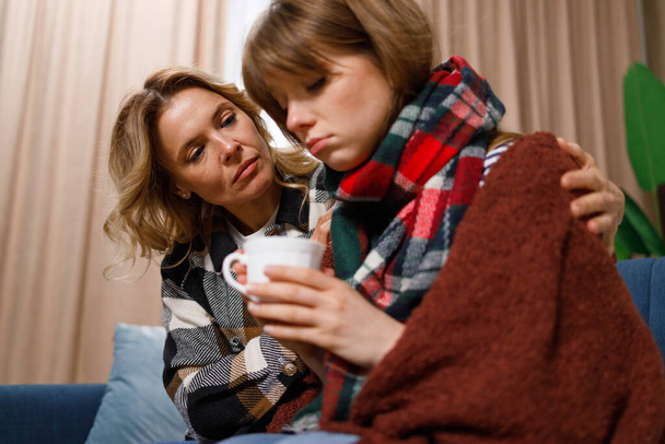 Mujer cariñosa abrazando a su hija que está enferma de gripe. Mala chica en una manta bebe té caliente mientras está sentada con su madre en el sofá en casa - Foto, imagen
