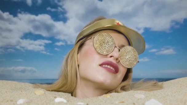 Egy csinos nő a fejéig eltemetve a parton napellenzővel és csillogó napszemüveggel. - Felvétel, videó