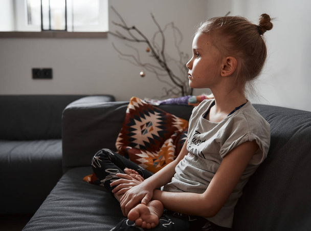 Schöne kleine Mädchen sitzt auf dem gemütlichen Sofa in Lotusposition im Wohnzimmer zu Hause und schaut fern - Foto, Bild