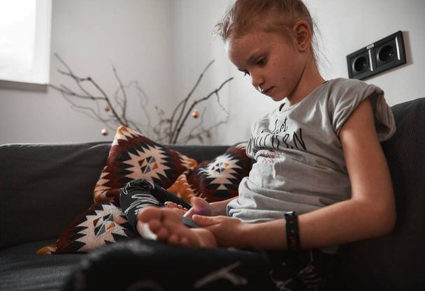 Cute child girl sitting on sofa in living room use smartphone and play online games. Holding phone and looking on screen device. Kid sitting on the couch and rest in living room. Technology concept. - Photo, Image