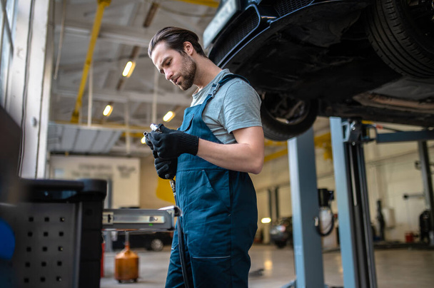 Serious focused young Caucasian automobile mechanic looking at the drill and nozzle in his hands - Photo, image