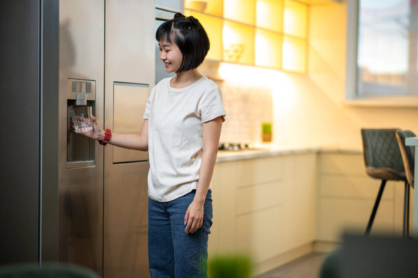 Smiling contented young woman standing in the kitchen and putting a container underneath the refrigerator water dispenser - Photo, image