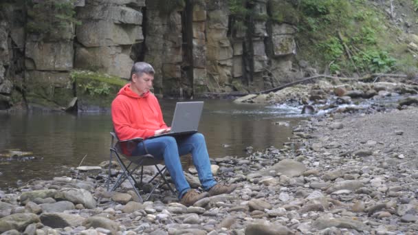 Человек работает на ноутбуке рядом с ручьем в природе. Концепция фрилансера, цифрового кочевника или удаленного офиса. 4K - Кадры, видео